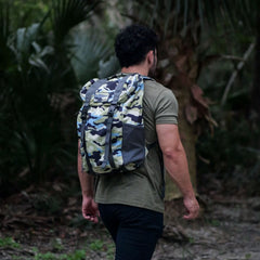 In a dense forest, a person is seen wearing a green t-shirt and black pants, carrying the M24 - Cordura by GORUCK. This modern rucksack, crafted with durable materials and featuring a camouflage-patterned design, seamlessly blends into the lush surroundings. Its bombproof laptop compartment guarantees safety on their adventurous journey.