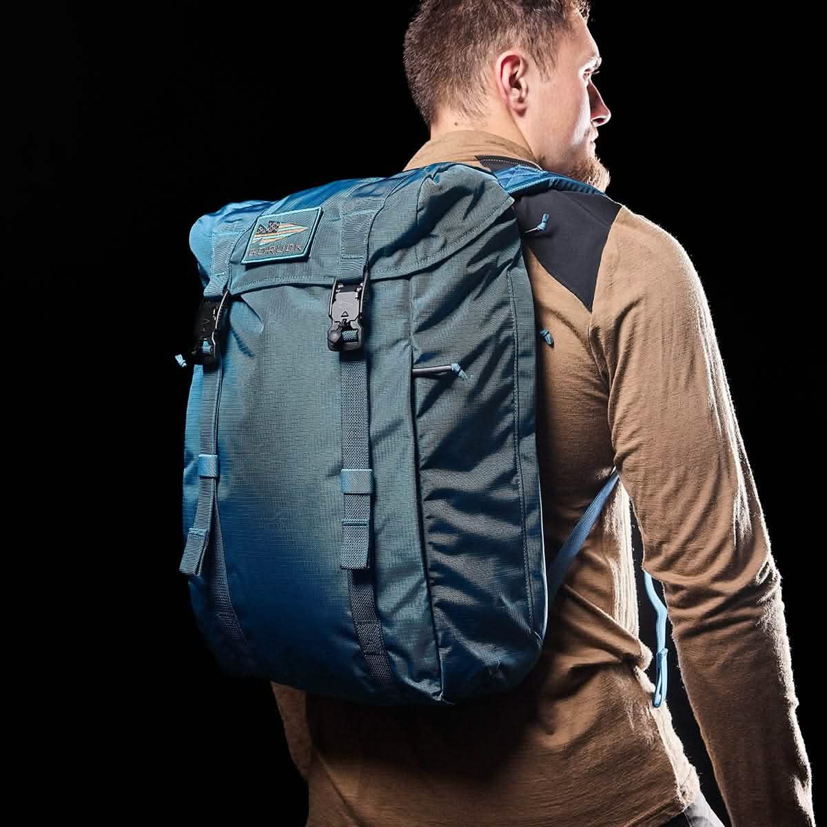 A man with short hair is wearing a tan long-sleeve shirt and carrying a Tidal Blue M23 from GORUCK. The pack features Ripstop ROBIC® material and has multiple straps. He is looking to the side against a black background.