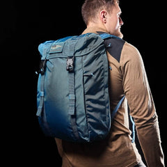 A man with short hair is wearing a tan long-sleeve shirt and carrying a Tidal Blue M23 from GORUCK. The pack features Ripstop ROBIC® material and has multiple straps. He is looking to the side against a black background.