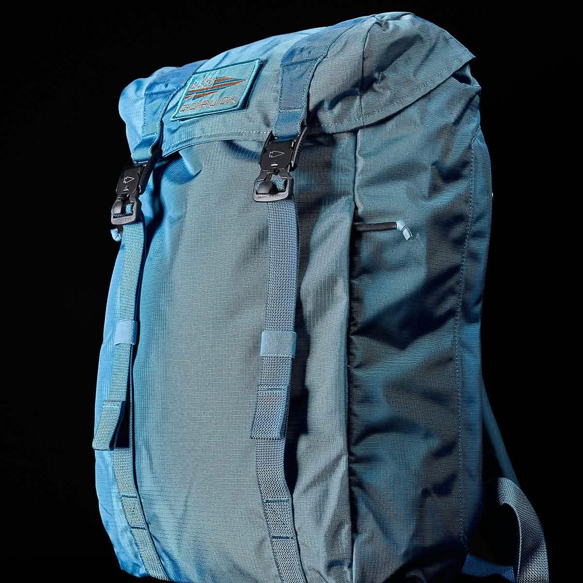 The GORUCK M23 - Ripstop ROBIC® rucksack in Tidal Blue and Speed Tan features black buckles and multiple compartments against a black background. Its durable fabric, reminiscent of a military backpack, is designed for outdoor use with a top flap and several adjustable straps.