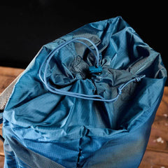 A close-up of the GORUCK M23 military backpack in Tidal Blue and Speed Tan, with its top drawstring closure open, revealing the fabric and cords. The 21L rucksack rests on a wooden surface against a dark background.