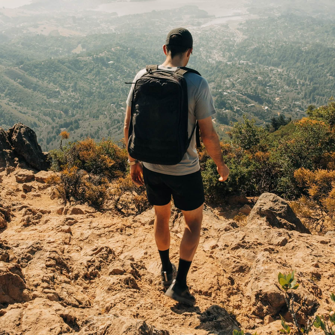 A person wearing a cap, T-shirt, shorts, and a pair of GORUCK's Mackall - Black + Forged Iron shoes is hiking down a rocky trail. They overlook a scenic view of a town surrounded by trees and hills under a clear sky. The trek feels like Special Forces training, pushing them beyond the ordinary with each step.