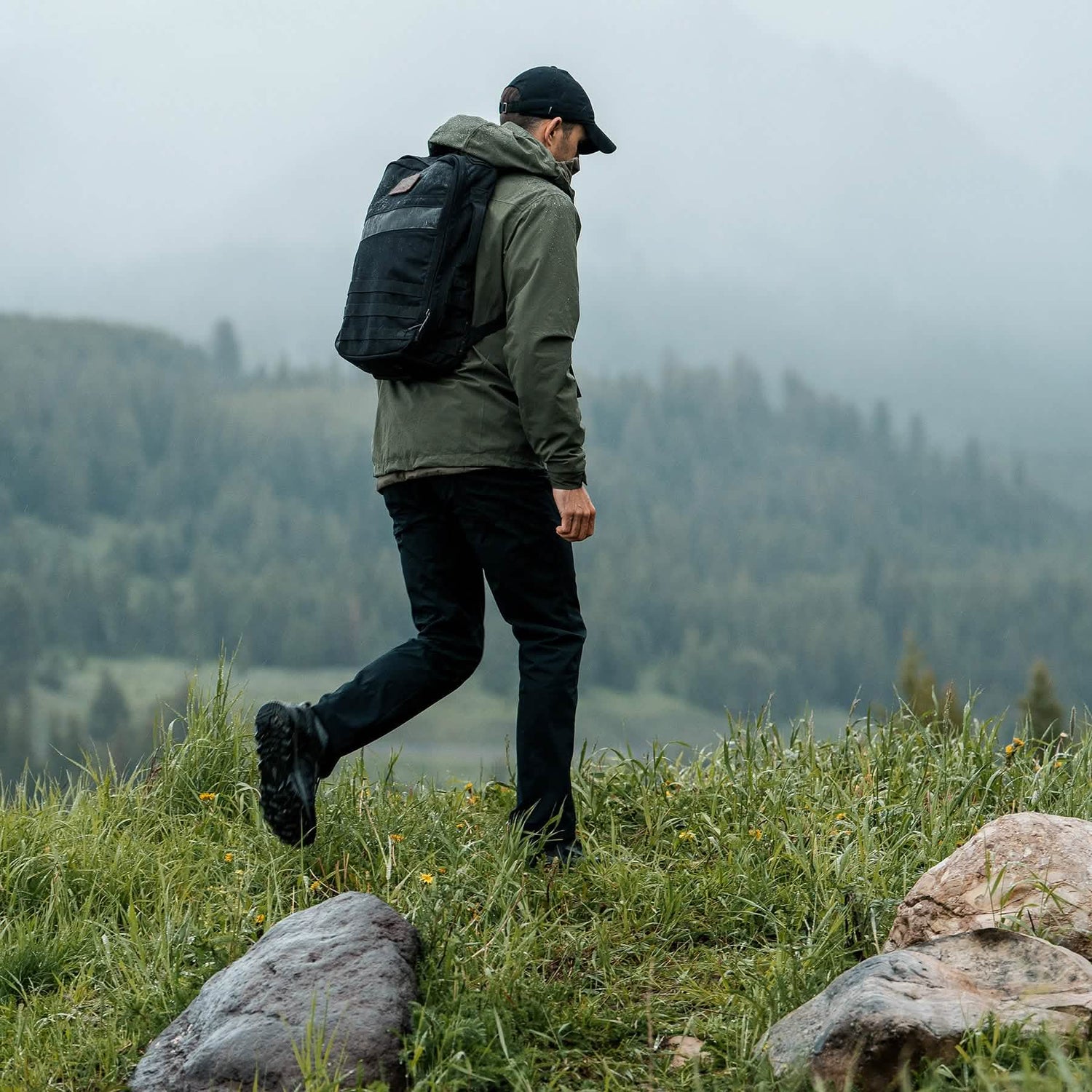A man, equipped with trail shoes, wearing a green Mackall jacket by GORUCK, black pants, and a backpack walks on a grassy hilltop with misty mountains and a forest in the background. He is facing away, heading toward the scenic landscape.