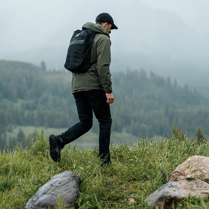 A man, equipped with trail shoes, wearing a green Mackall jacket by GORUCK, black pants, and a backpack walks on a grassy hilltop with misty mountains and a forest in the background. He is facing away, heading toward the scenic landscape.
