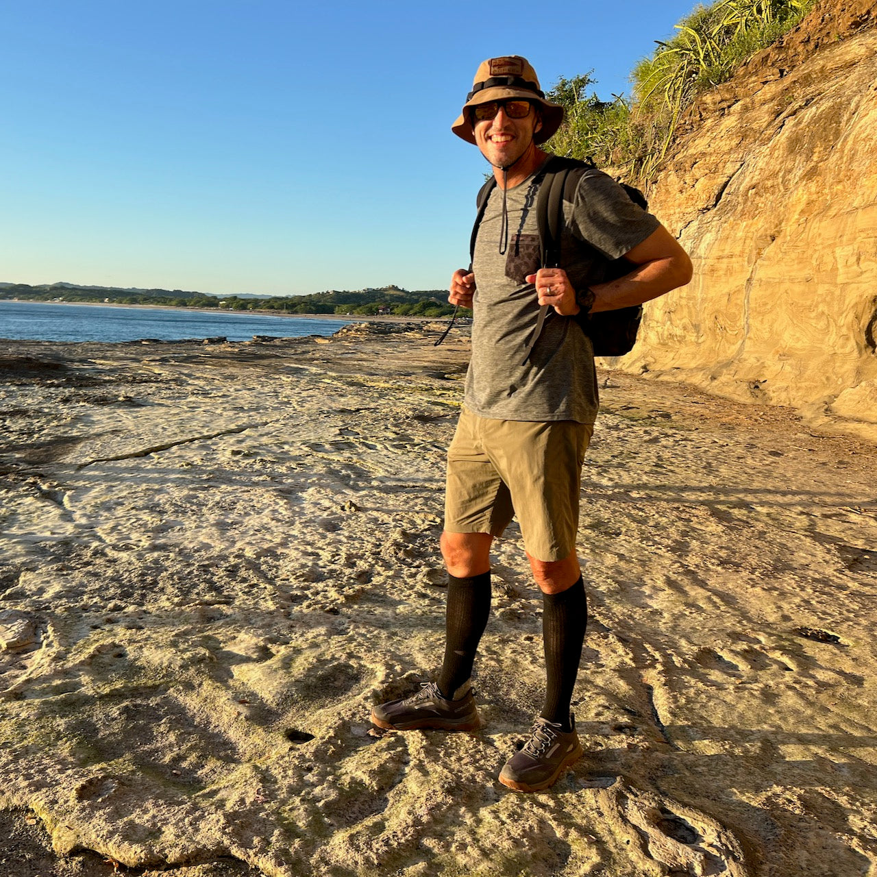 Man wearing Mackall – Forged Iron + Chiseled Stone + Gum trail shoes and backpack smiles on a sunlit rocky coastline, ready for outdoor adventures.