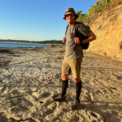 Man wearing Mackall – Forged Iron + Chiseled Stone + Gum trail shoes and backpack smiles on a sunlit rocky coastline, ready for outdoor adventures.