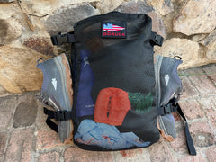 The Mackall - Forged Iron + Chiseled Stone + Gum all-terrain rucking shoes are attached to a black GORUCK backpack, resting against a stone wall on a brick surface.