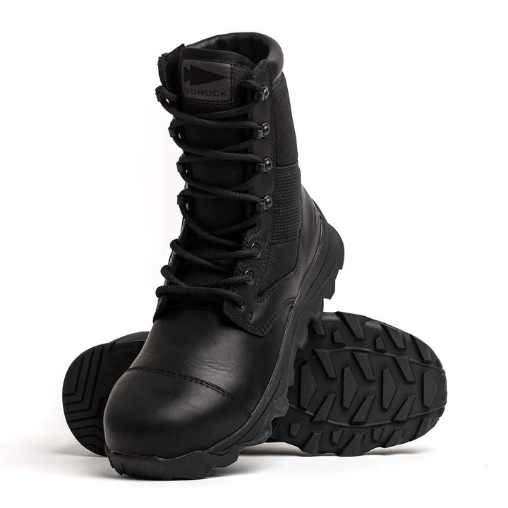 The MACV-2 Safety Boot - High Top, crafted by GORUCK to meet Special Forces standards, features rugged soles. One boot stands upright, while the other lies on its side, showcasing the thick tread pattern. The brand logo is visible on the tongue against a plain white background.