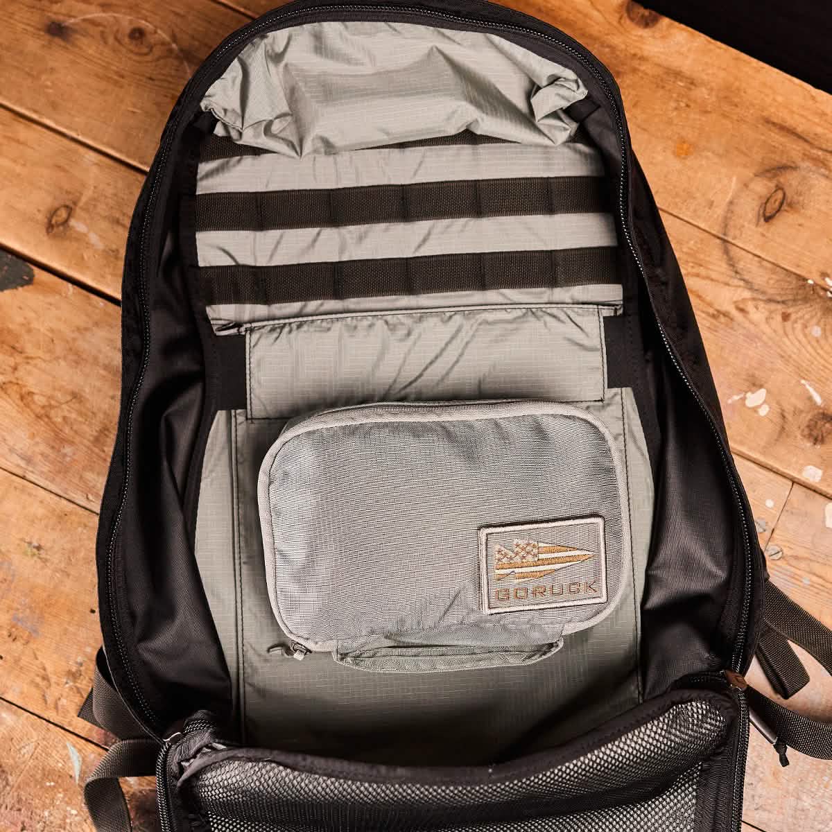 An open backpack on a wooden floor reveals its rucksack organization. Crafted from 420D ROBIC Nylon, the black bag's interior features GORUCK's Shadow Pockets - Ripstop ROBIC® for efficiency. Inside, a small gray pouch with a GORUCK patch and flag logo is neatly attached.
