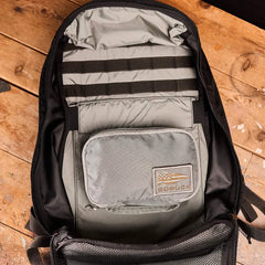 An open backpack on a wooden floor reveals its rucksack organization. Crafted from 420D ROBIC Nylon, the black bag's interior features GORUCK's Shadow Pockets - Ripstop ROBIC® for efficiency. Inside, a small gray pouch with a GORUCK patch and flag logo is neatly attached.