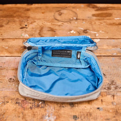 A Shadow Pocket from GORUCK, featuring an open design in light grey and blue, crafted from 420D Ripstop ROBIC® Nylon, sits on a wooden surface. Perfect for organizing within your rucksack, its blue interior includes a mesh pocket. The lid displays a black tag with text, highlighting the utility of the Shadow Pockets collection.