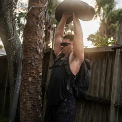 Man wearing black sleeveless performance tank and backpack lifting weighted sandbag overhead outdoors
