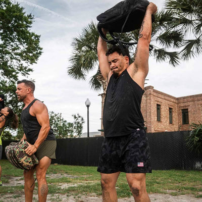 Two men enjoy exercising outdoors with one lifting a backpack over his head and the other holding a bag at waist height. Clad in GORUCK Men's Performance Tanks - ToughMesh and black athletic shorts, they revel in the fresh air surrounded by trees and a building in the background.