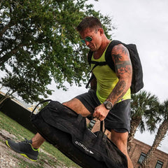 Athletic man wearing acid lime tank top and backpack lifting heavy black GORUCK bag outdoors