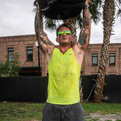 Man with tattoos wearing acid lime performance tank and sunglasses lifting heavy bag outdoors with palm trees