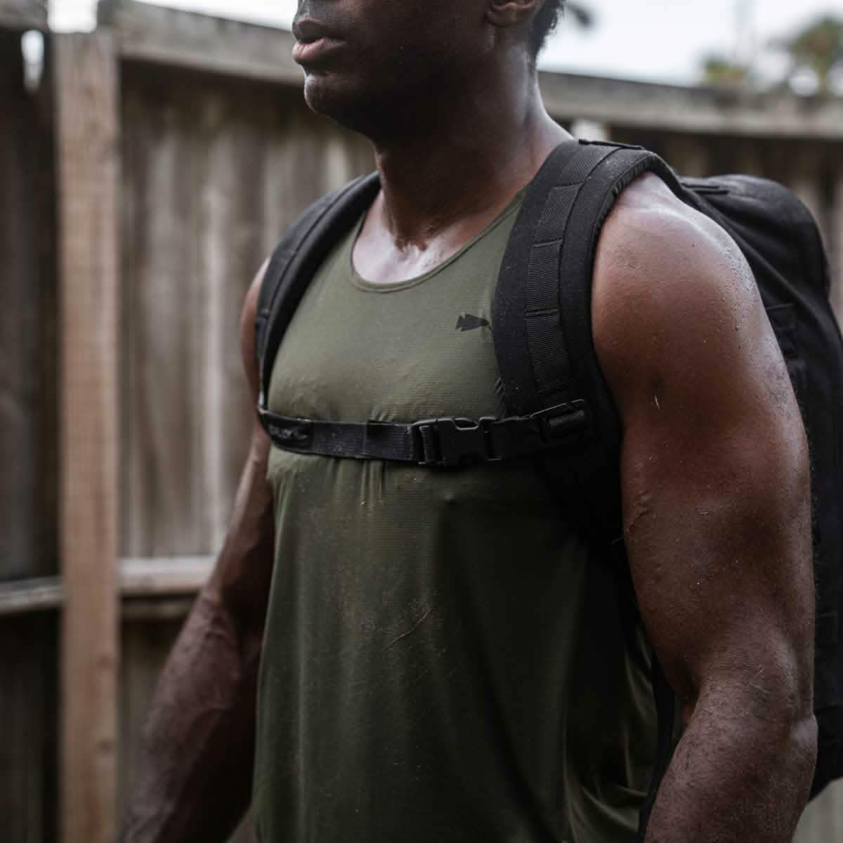 A man in a Men's Performance Tank - ToughMesh and a backpack stands outdoors near a wooden fence.