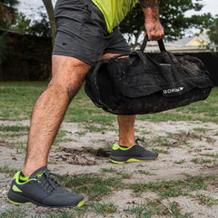 Man wearing GORUCK shorts and shoes lifting a black GORUCK rucking bag outdoors on grass