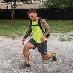 Fit man with tattoos in neon tank top and shorts performing lunge exercise outdoors wearing a backpack