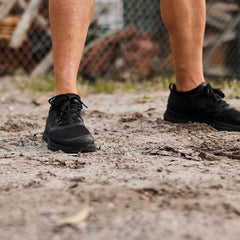 Close-up of a person wearing black GORUCK men's Rough Runners standing on dirt ground with a chain-link fence background