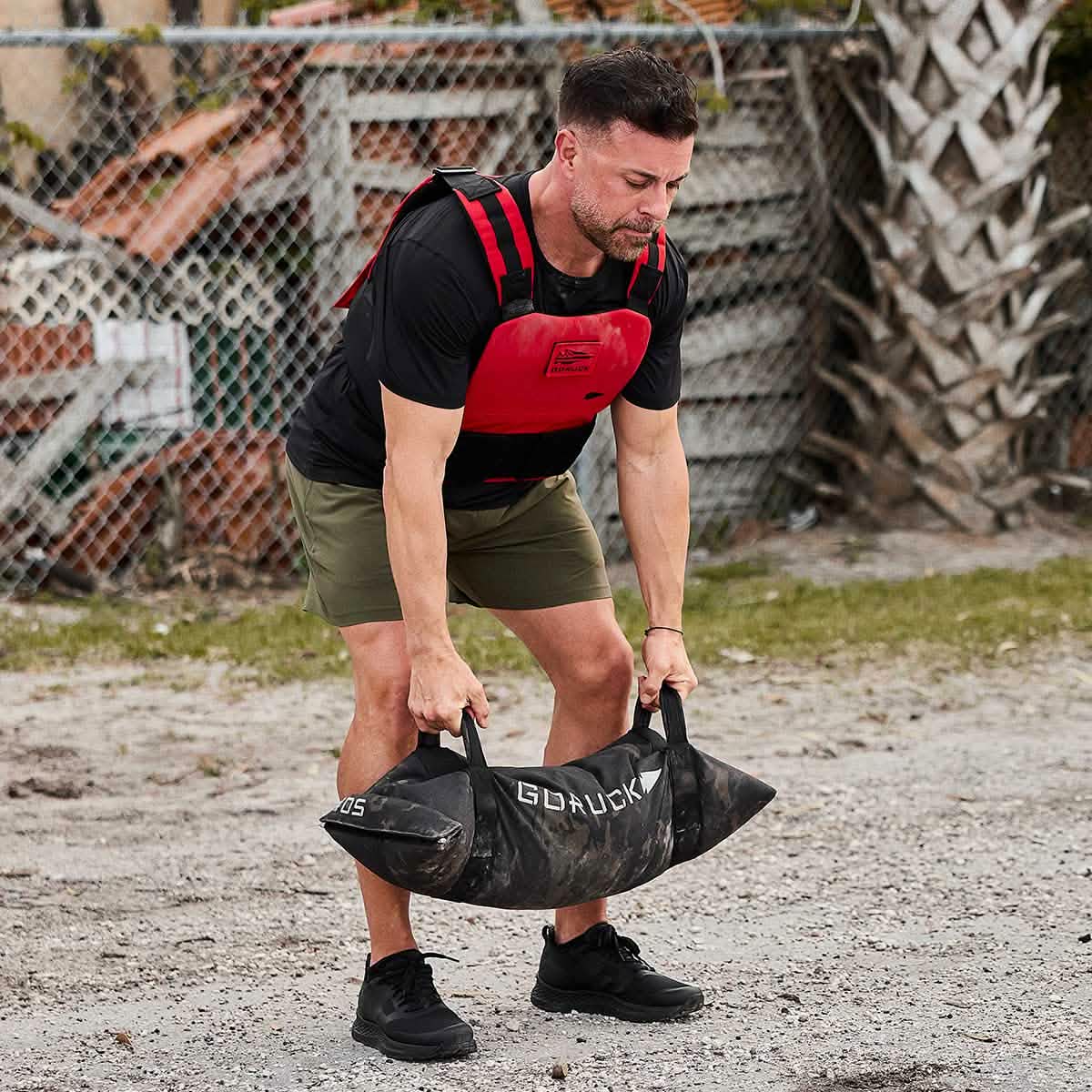 A man wearing the Men's Rough Runner - Blackout weighted vest from GORUCK pairs it with green shorts and lightweight running shoes, as he hoists a black sandbag branded with GORUCK. He's outside on a dirt and gravel terrain, surrounded by a chain-link fence and foliage, capturing the essence of a Rough Runner challenge.