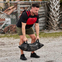 Man wearing red weighted vest and black shoes lifting a heavy black GORUCK sandbag outdoors on gravel
