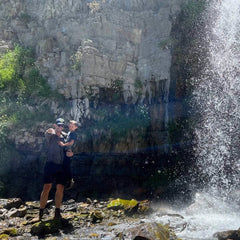 A man wearing GORUCK's Merino Challenge Socks - Crew stands near a waterfall, holding a child as sunlight filters through the spray. Surrounded by rocky terrain and patches of greenery, they enjoy the moment with confidence in their reliable footwear backed by Scars Lifetime Guarantee's promise.