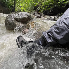 A person wearing gray pants and black shoes, complemented by cozy Merino Challenge Socks - Crew from GORUCK, is standing in a flowing stream. Water rushes over their foot, surrounded by rocks and lush, green foliage.