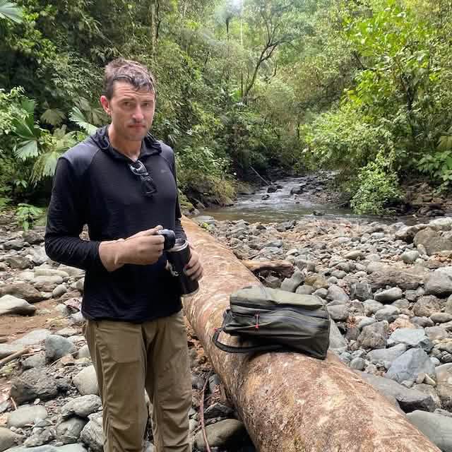 A person, wearing a Men’s Commando Pullover - Merino Wool from GORUCK, stands on a rocky streambed in a forest, holding a canteen. Their backpack rests on a large fallen log beside them. The area is lush with greenery and dense vegetation.