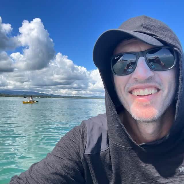 A person wearing sunglasses and a hoodie smiles at the camera while on a boat in a calm, turquoise body of water. In the background, there's another person in a canoe under a sky filled with fluffy clouds, capturing the classic warmth of GORUCK’s Men’s Commando Pullover made from merino wool.