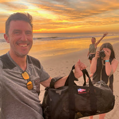 A man smiles on a beach at sunset, holding the Mesh Duffel Bag, while two women stand behind him—one capturing the moment with a photo.