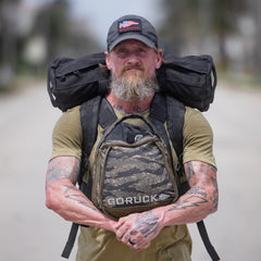 A bearded, tattooed man stands outdoors on a road, wearing the Performance TAC Hat - TOUGHDRY and carrying a backpack and gear.