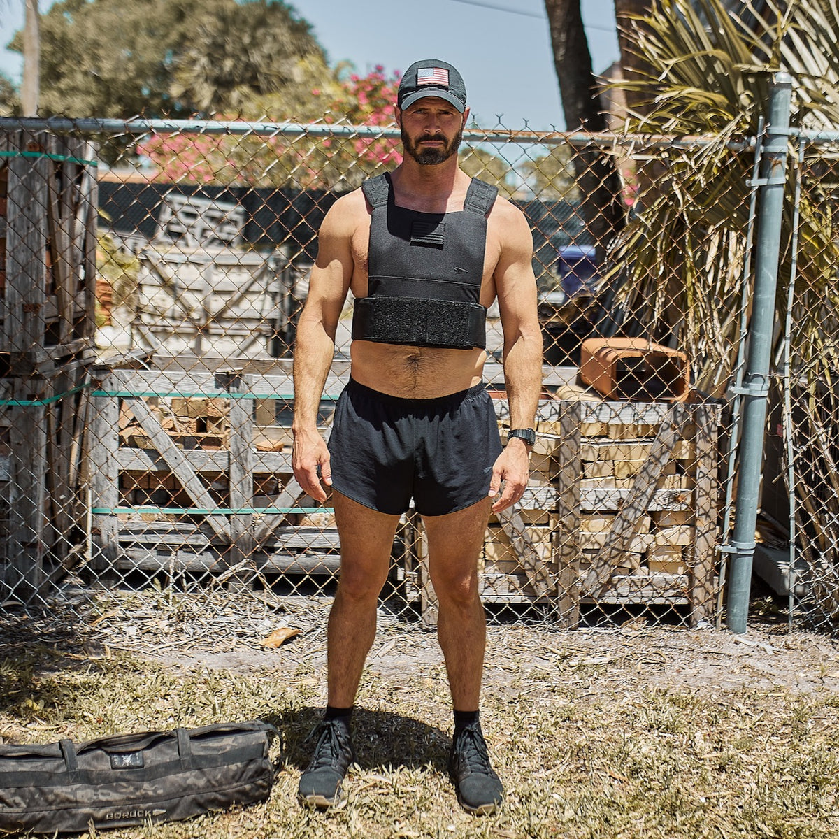A man in black workout gear and a cap stands outdoors by a chain-link fence on a sunny day, wearing the Training Weight Vest - Black for added resistance during his workout.