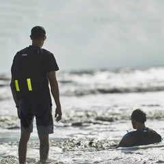 Two men with backpacks at the beach, one standing in shallow water, the other sitting in the surf with waves