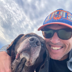 A man in sunglasses and a hat hugs a brown dog on a beach beneath a cloudy sky, both smiling at the camera—a heartwarming moment that embodies the unbreakable bond honored by the Monster Forever Tee - PREORDER and treasured by all who remember beloved companions across the Rainbow Bridge.