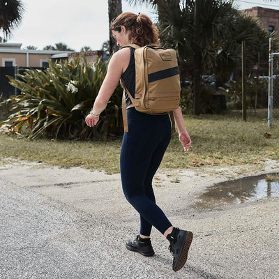 A person with red hair walks outdoors on a cloudy day, sporting a tan backpack and GORUCK Women’s Training Leggings Pocket made from ToughFlex fabric. They stride confidently in sneakers along a paved path, flanked by lush greenery and a chain-link fence.