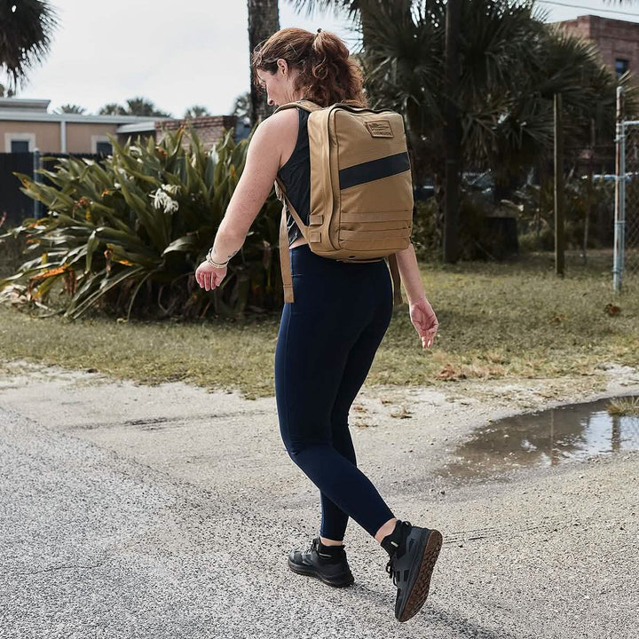 A person with red hair walks outdoors on a cloudy day, sporting a tan backpack and GORUCK Women’s Training Leggings Pocket made from ToughFlex fabric. They stride confidently in sneakers along a paved path, flanked by lush greenery and a chain-link fence.
