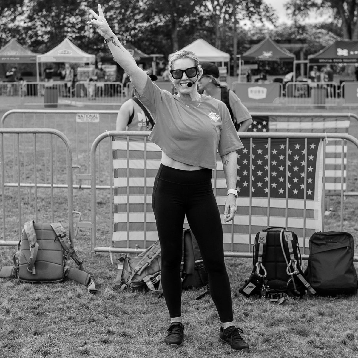 Woman in sunglasses and athletic wear smiles and raises a peace sign in front of an American flag at an outdoor event.