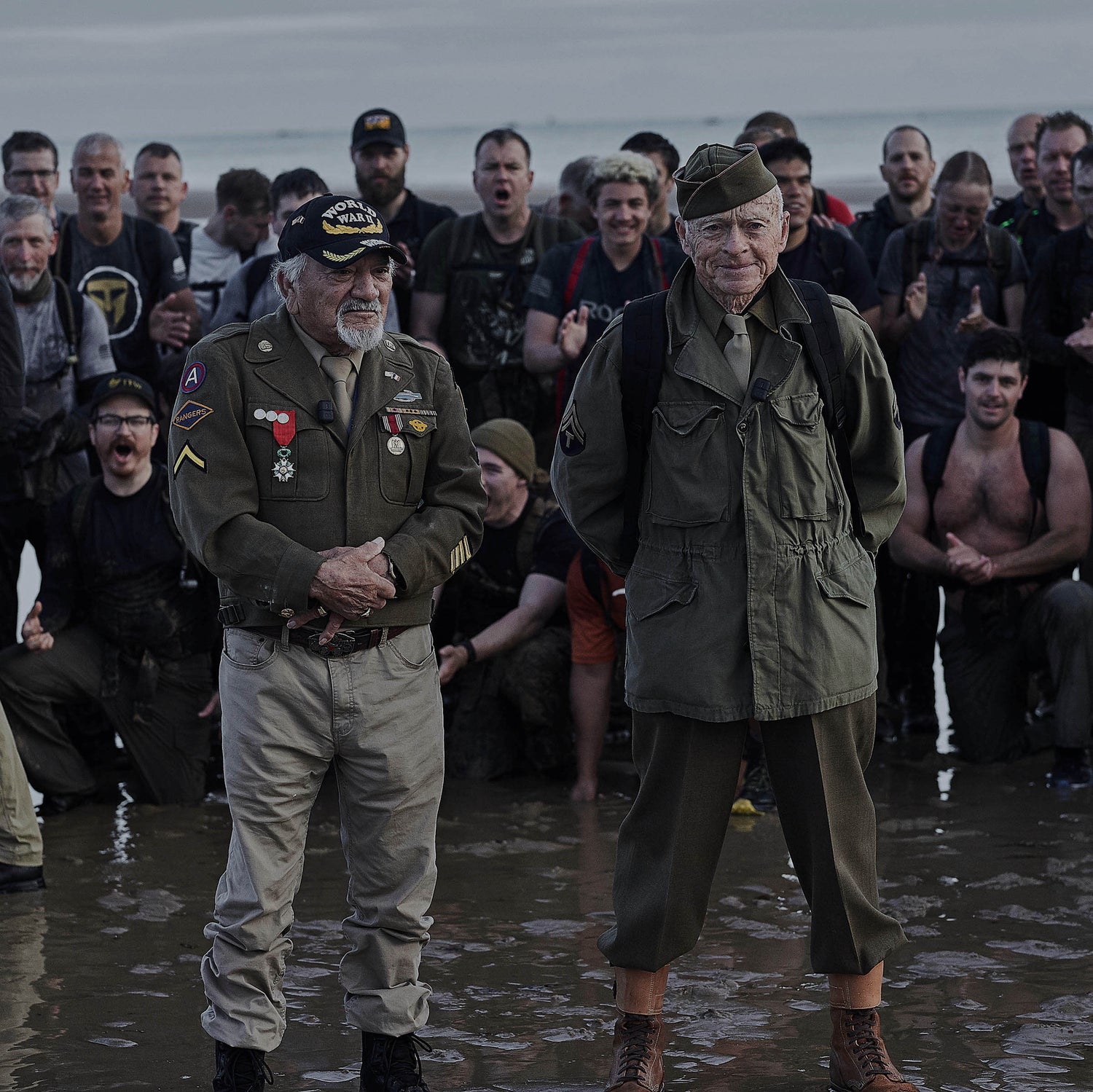Two elderly veterans stand on a beach, surrounded by a group of people applauding them.