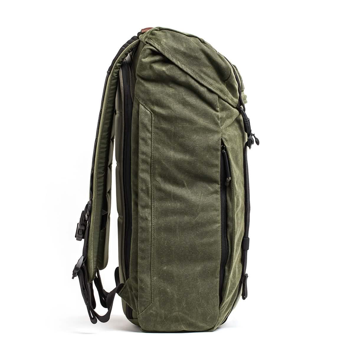 A side view of a green M23 - Waxed Canvas rucksack from GORUCK, featuring multiple zippers and adjustable shoulder straps, highlights its spacious and functional design. The waxed canvas material appears durable and ideal for various outdoor activities, consistent with GORUCK's standards.