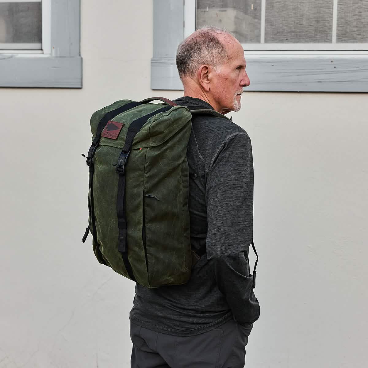A man in a dark long-sleeve shirt stands in profile, sporting the large, modern GORUCK M23 - Waxed Canvas rucksack. He is positioned in front of a light-colored wall that features two windows.