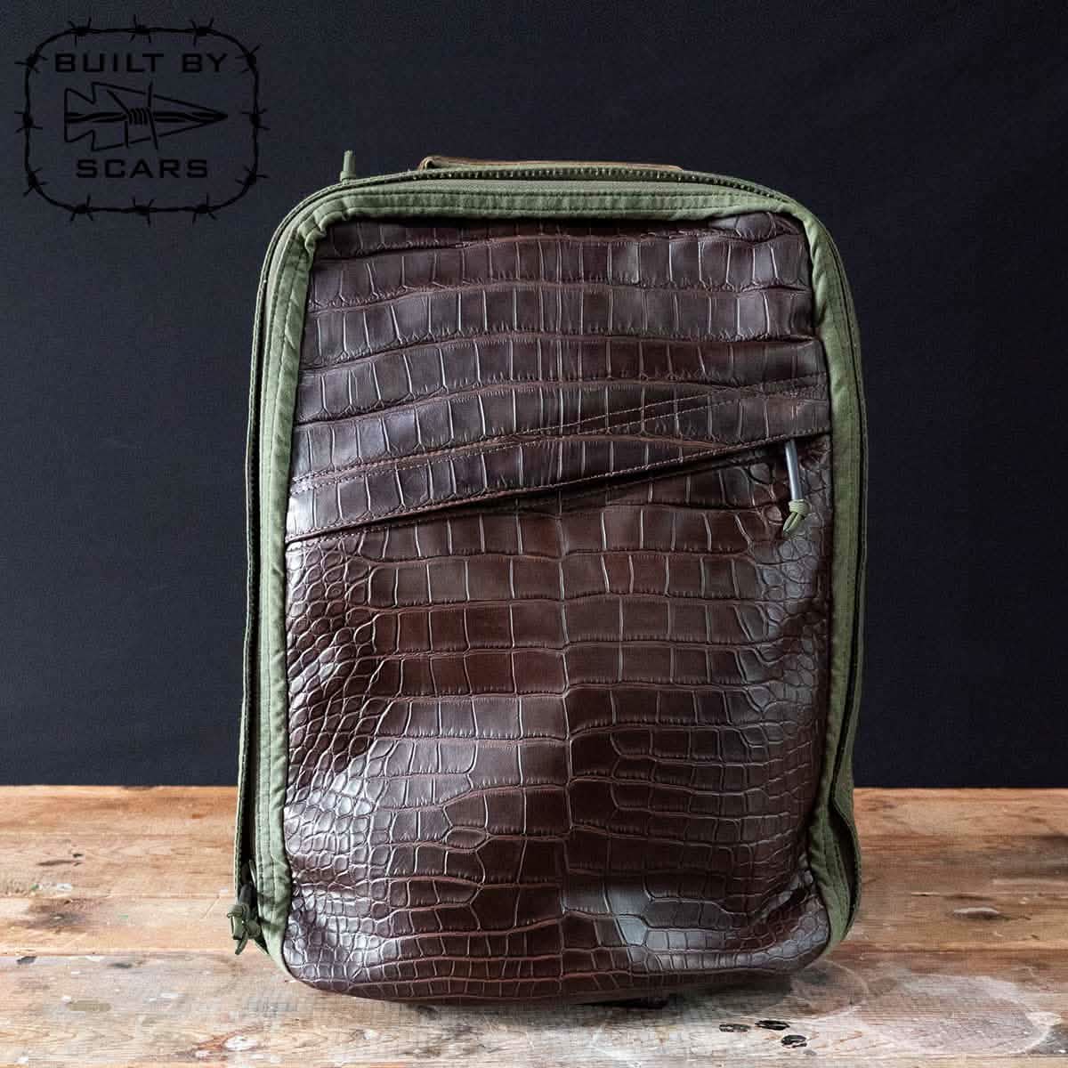 A GR1 Heritage backpack by GORUCK, featuring an alligator leather front panel in green, rests on a wooden table against a dark background. The top has a zip closure, and its design evokes the classic style of retro canvas rucksacks with the logo prominently displayed in the upper left corner.