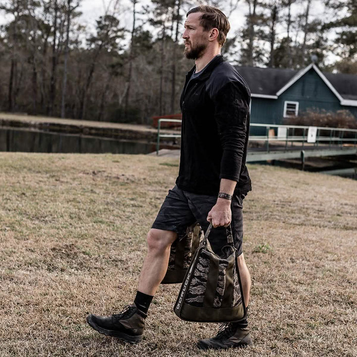 A man, clad in a black long-sleeve shirt and dark shorts, walks on grass near a pond carrying two camo-patterned bags. His GORUCK MACV-2 - Mid Top - Origins jungle boots crunch softly as he passes; behind him stands a green house with a white door, tucked among trees.