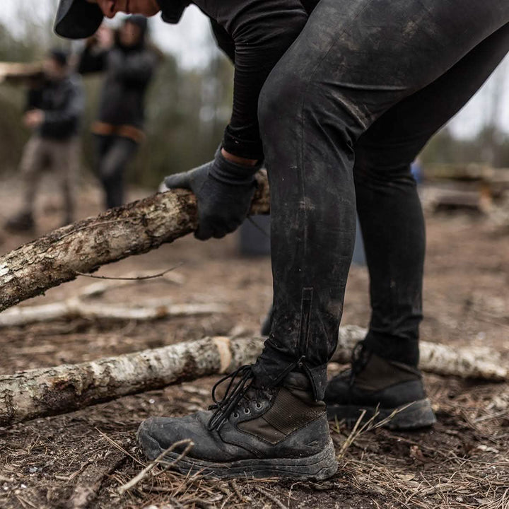A person dressed in rugged outdoor clothing, featuring the black MACV-2 - Mid Top - Origins boots from GORUCK and gloves, is seen bending over while holding a thick branch on a forest floor. Blurred figures and trees create a wilderness setting reminiscent of Special Forces operations in the background.