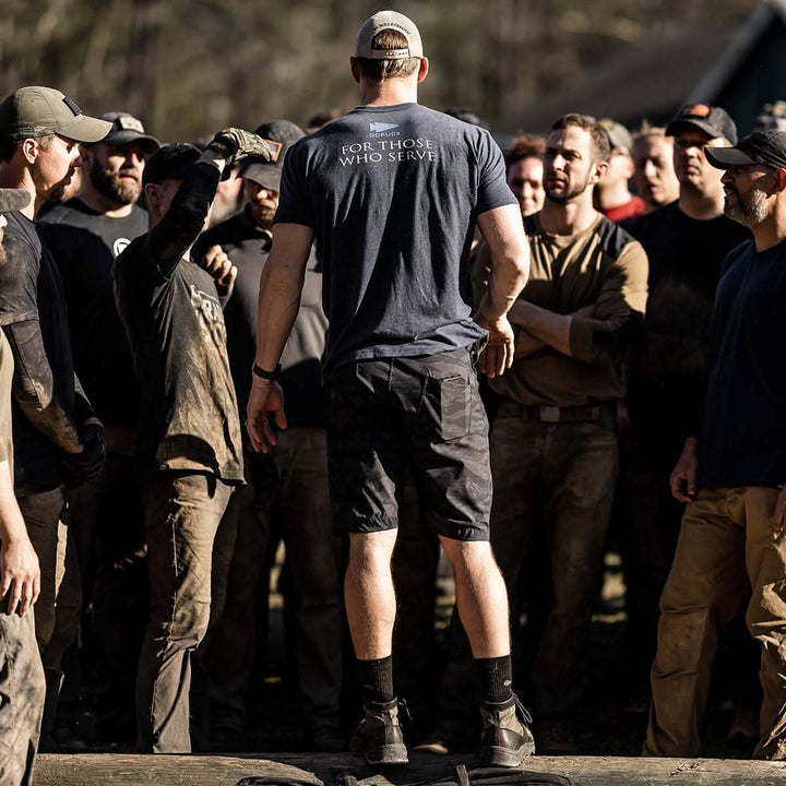 A group of men gathered outdoors, with one man in focus, wearing a shirt that says "For Those Who Serve" and sporting GORUCK's MACV-2 Mid Top Origins jungle boots. He stands on a log, surrounded by the men, some dressed in work attire reminiscent of Special Forces, in a sunlit area.