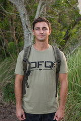 Certainly! Here’s a rewritten version using your product data: A man with short brown hair stands outdoors surrounded by lush greenery, sporting a khaki DFQ Tee - Tri-BlendX and a backpack. The bold “Don’t F*ing Quit” slogan on his shirt gives his adventurous look an extra boost of motivation.