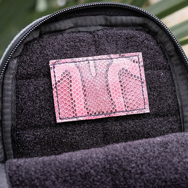 A close-up view of the Everglades Mystery Leather Patch by GORUCK reveals a stitched rectangular patch with a pink snakeskin pattern on a black fabric surface, creating a striking contrast with the darker background.