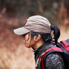 A person wearing a tan Performance TAC Hat - Slick - TOUGHDRY and a backpack, hiking outdoors in side profile, with a blurred natural background.