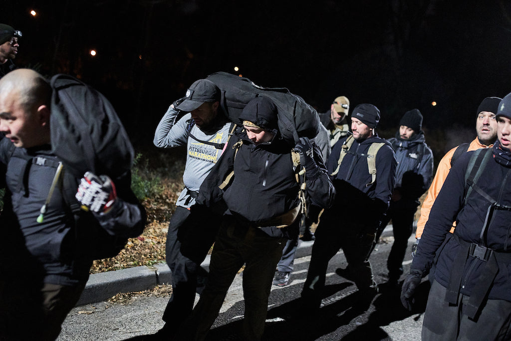 Group of people wearing outdoor gear carry heavy backpacks and a person during a nighttime endurance event.