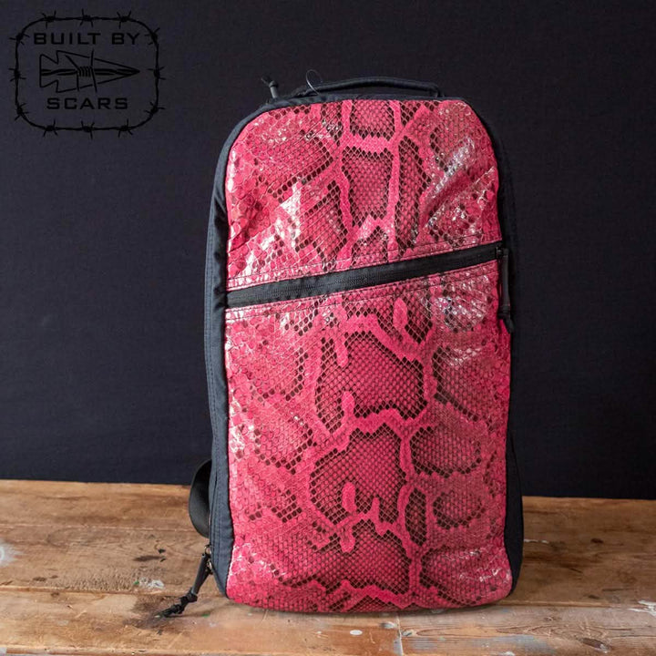 A Bullet Ruck Heritage - Pink Python backpack from GORUCK, featuring a red snake skin texture with black accents, is placed on a wooden surface, giving off a heritage vibe. The Built by Scars logo prominently appears in the top left corner against a black background, evoking the rugged elegance synonymous with waxed canvas designs.