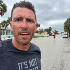 Man wearing the "It's Not About You - Tri-BlendX - Unisex" shirt takes a selfie on a wet, palm-lined street with houses in the background; another person appears in the distance. Printed in the USA.